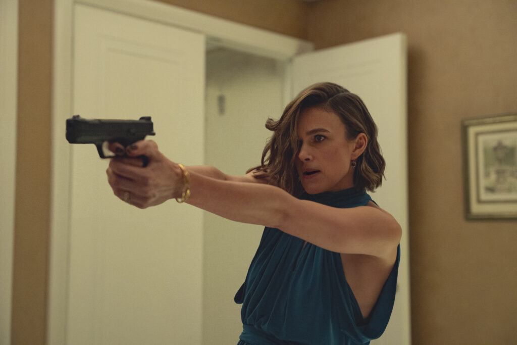 "Woman in blue dress aiming a handgun with both hands in a tense indoor setting, standing by a white door with a picture frame on the beige wall in the background."